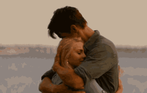 A Man Is Holding A Woman In His Arms In Front Of A Body Of Water . GIF