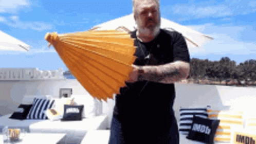 A Man Is Holding A Yellow Umbrella In Front Of A Couch With A Pillow That Says Imdb GIF