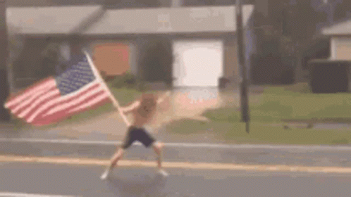 A Man Is Holding An American Flag While Walking Down A Street . GIF