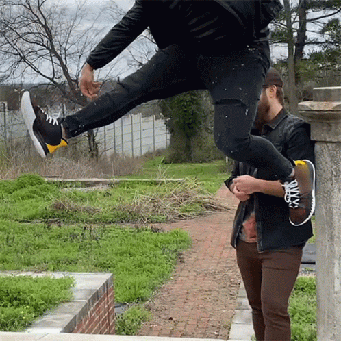 A Man Is Holding Another Man 'S Leg While He Jumps Over A Brick Wall GIF