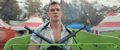A Man Is Holding Two Guns On His Shoulders While Riding A Green Ferris Wheel At A Carnival . GIF