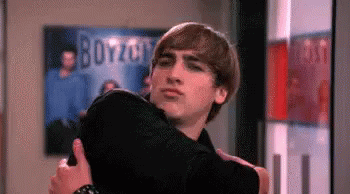 A Man Is Hugging A Woman In A Room With A Poster On The Wall That Says Boyzoo . GIF