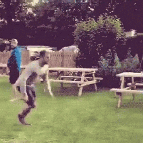 A Man Is Jumping In The Air In A Field With Picnic Tables In The Background GIF
