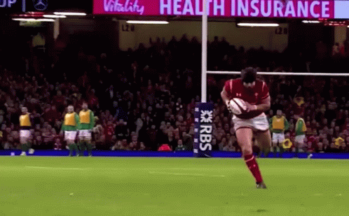 A Man Is Kicking A Rugby Ball On A Field With A Sign That Says Health Insurance In The Background . GIF