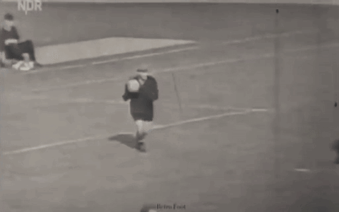 A Man Is Kicking A Soccer Ball On A Field While Another Man Sits On The Ground . GIF