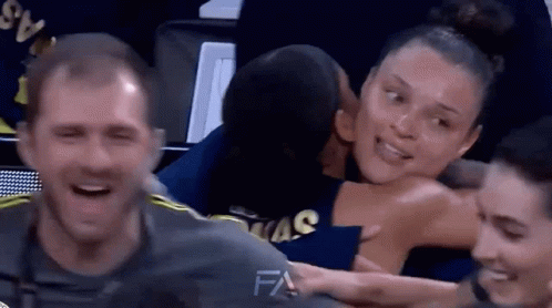 A Man Is Kissing A Woman On The Cheek While Sitting In A Basketball Stadium . GIF