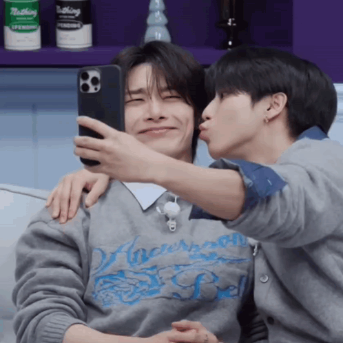 A Man Is Kissing Another Man On The Cheek While They Take A Selfie GIF