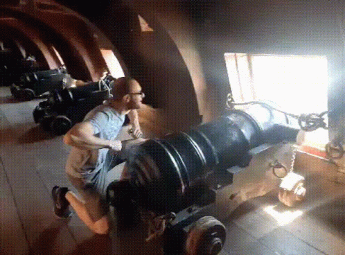 A Man Is Kneeling Down In Front Of A Cannon With A Window In The Background GIF