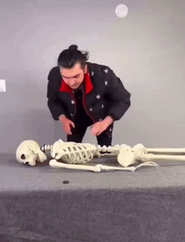 A Man Is Kneeling Down Next To A Skeleton And A Skull On A Table . GIF