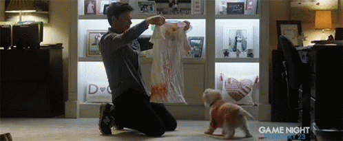 A Man Is Kneeling Down Next To A Small White Dog Wearing A Red Shirt That Says " Game Night " On It GIF