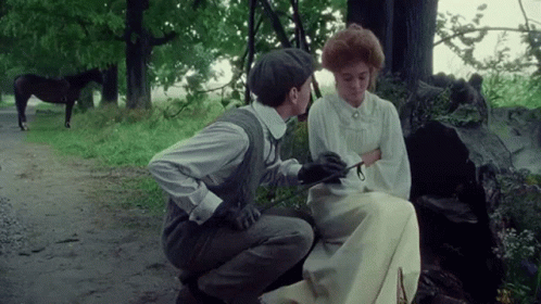 A Man Is Kneeling Down Next To A Woman Sitting On A Bench With A Horse In The Background . GIF