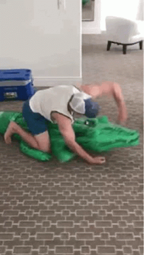A Man Is Kneeling On The Floor With A Green Inflatable Alligator . GIF