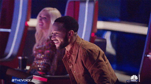 A Man Is Laughing While Sitting In Front Of A Nbc Sign GIF