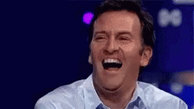 A Man Is Laughing With His Mouth Open While Sitting On A Stage . GIF