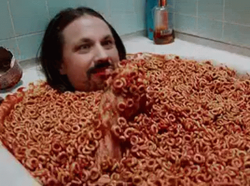 A Man Is Laying In A Bathtub Filled With Cereal Rings . GIF