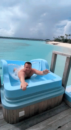 A Man Is Laying In A Blue Tub On A Dock Overlooking The Ocean GIF