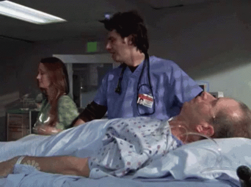 A Man Is Laying In A Hospital Bed While A Nurse Stands Behind Him . GIF