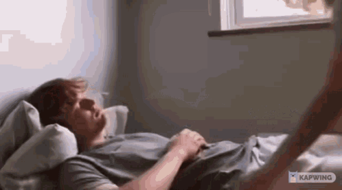 A Man Is Laying On A Bed Reading A Book And Looking Out A Window . GIF