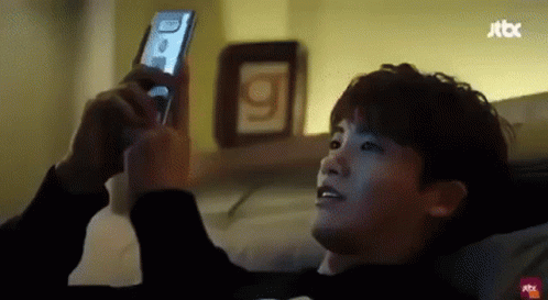 A Man Is Laying On A Bed Using A Cell Phone . GIF
