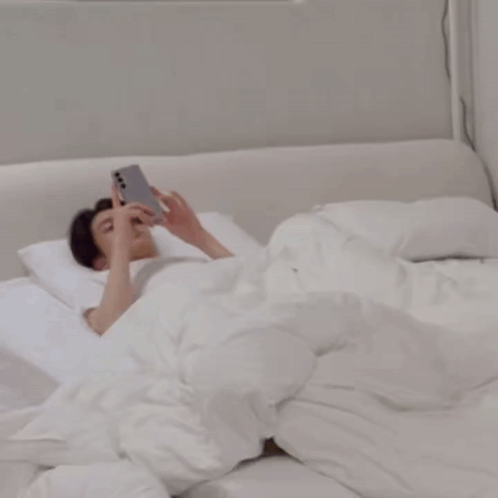 A Man Is Laying On A Bed While Another Man Looks At His Phone GIF