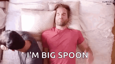 A Man Is Laying On A Bed With A Pillow On His Head And Another Man Is Standing Behind Him . GIF