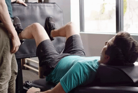 A Man Is Laying On A Bench In A Gym While A Man Stands Behind Him . GIF