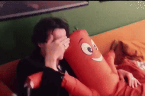 A Man Is Laying On A Couch Holding A Stuffed Sausage GIF