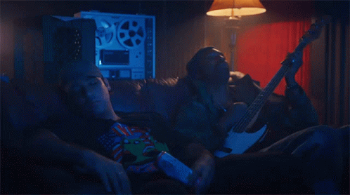 A Man Is Laying On A Couch While Another Man Holds A Guitar GIF