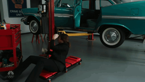 A Man Is Laying On A Rolling Chair In Front Of A Car That Has A Sign That Says Crime Scene Unit On It GIF