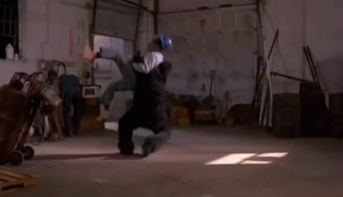A Man Is Laying On His Back In A Garage While Another Man Looks On . GIF