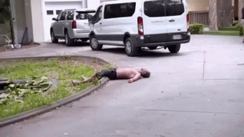 A Man Is Laying On The Ground In A Driveway Next To A White Van . GIF