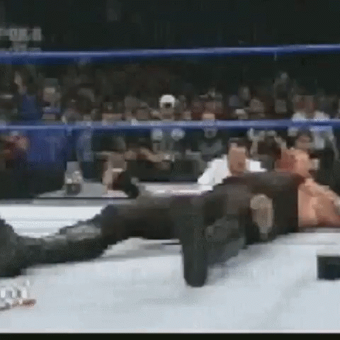 A Man Is Laying On The Ground In A Wrestling Ring While A Crowd Watches . GIF