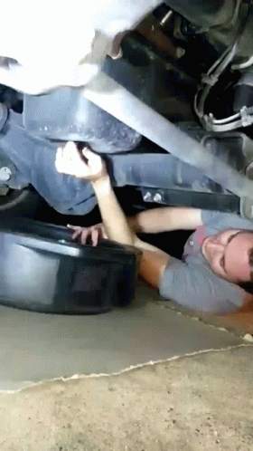 A Man Is Laying On The Ground Under A Car With A Black Bucket In Front Of Him GIF