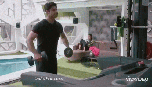 A Man Is Lifting A Dumbbell In A Living Room While A Woman Sits On A Couch . GIF