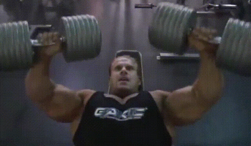 A Man Is Lifting Dumbbells In A Gym Wearing A Black Tank Top That Says Fake GIF