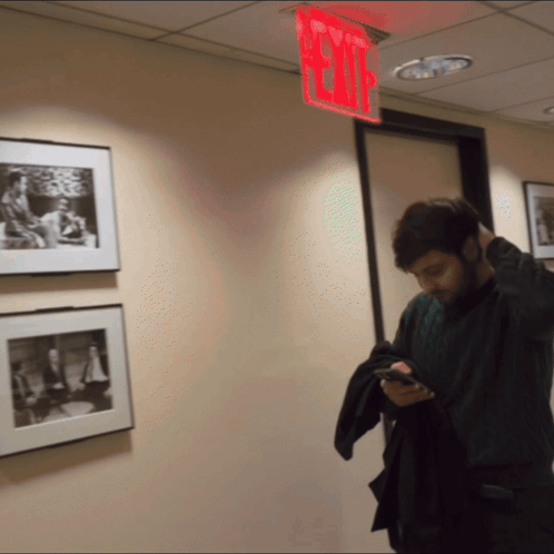 a man is looking at his phone in a hallway with a red exit sign above him