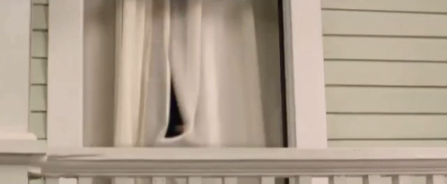 A Man Is Looking Out Of A Window With Curtains . GIF