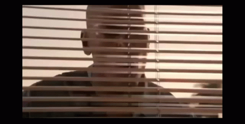 A Man Is Looking Through A Window With Blinds . GIF