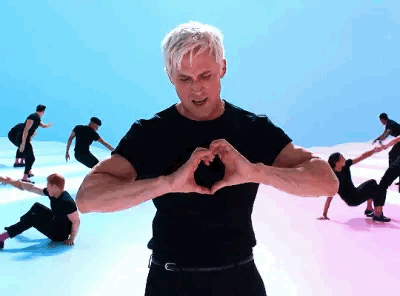 A Man Is Making A Heart Shape With His Hands While A Group Of People Are Dancing In The Background . GIF