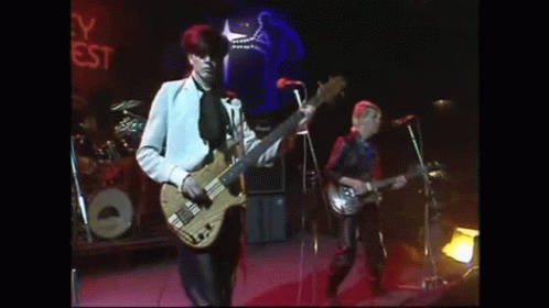 A Man Is Playing A Bass Guitar On A Stage While Another Man Sings Into A Microphone . GIF