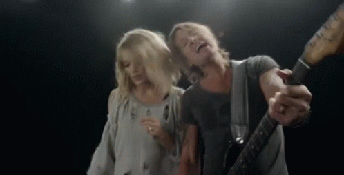 A Man Is Playing A Guitar And Singing Into A Microphone While A Woman Stands Behind Him . GIF