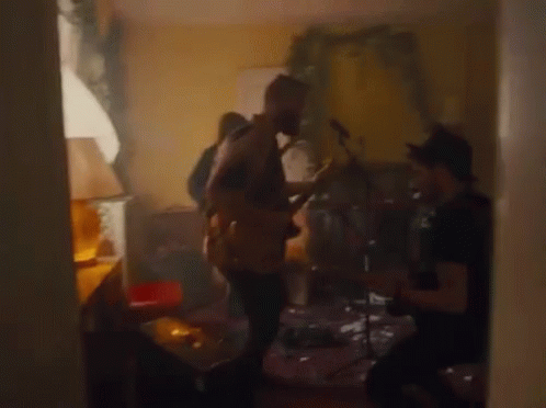 A Man Is Playing A Guitar In A Living Room While Another Man Plays Drums . GIF