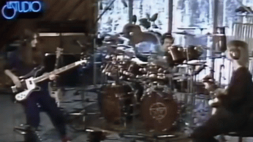 A Man Is Playing A Guitar In Front Of A Drum Set With A Neon Sign That Says Studio GIF