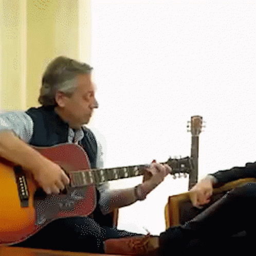 a man is playing an acoustic guitar while another man sits in a chair