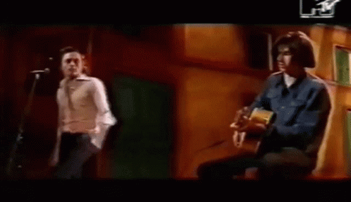 A Man Is Playing An Acoustic Guitar While Another Man Stands Behind Him . GIF