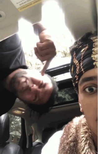 A Man Is Pointing At Another Man 'S Face In A Car With A Warning Sign On The Visor GIF
