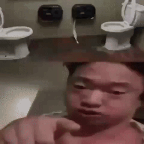 A Man Is Pointing At The Camera In A Bathroom With Toilets Behind Him . GIF