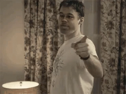 A Man Is Pointing At The Camera While Standing In Front Of A Lamp In A Room . GIF