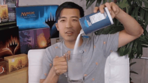 A Man Is Pouring Milk Into A Glass In Front Of Boxes Of Magic The Gathering Cards GIF