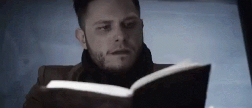 A Man Is Reading A Book With A Scarf Around His Neck . GIF